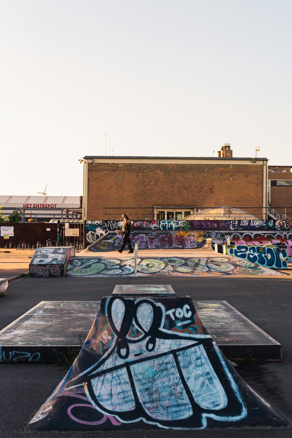 Una persona lleva una patineta por un parque de patinaje urbano cubierto de grafitis.