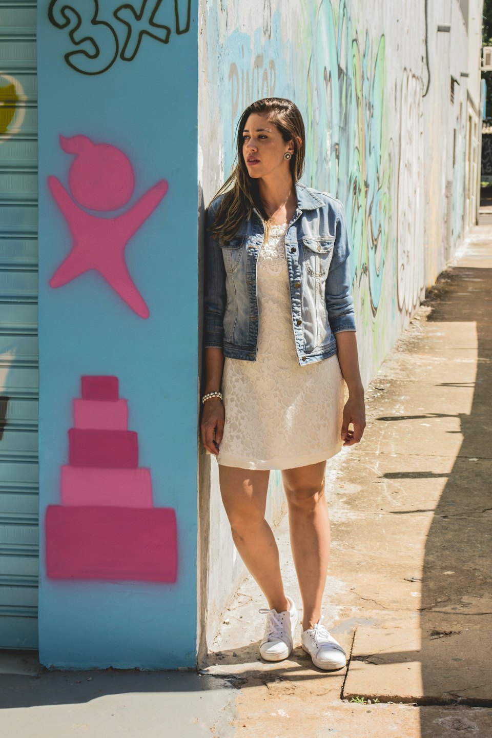 Mujer con vestido blanco y chaqueta vaquera en callejón con grafitis.